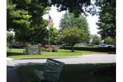Outdoor view of a landscaped area with green grass, trees, a flagpole with an American flag, a sign for Lebanon Valley Home, and a parking lot in the background. There is a green bench in the foreground under the shade of trees.
