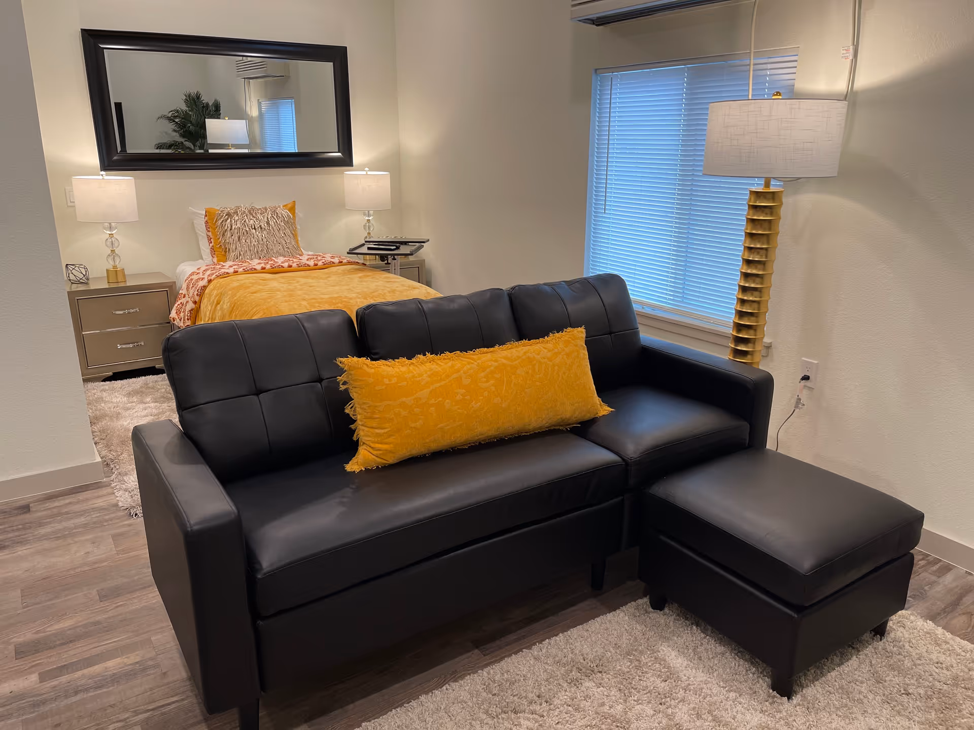 A cozy room featuring a black leather sofa with a yellow decorative pillow and matching ottoman on a light-colored rug. Behind the sofa is a bed with yellow and patterned bedding, flanked by two bedside tables each with a lamp. A large rectangular mirror hangs on the wall above the bed, and a tall gold floor lamp stands near a window with closed blinds.