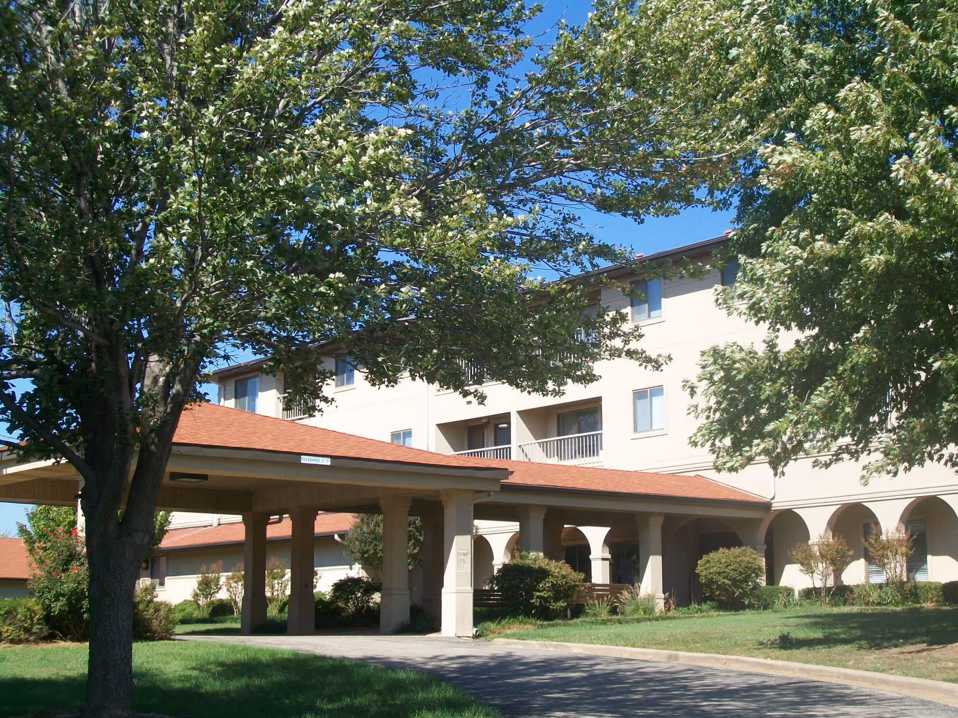 Exterior view of a multi-story senior living facility building with a covered entrance, surrounded by green trees and bushes under a clear blue sky.