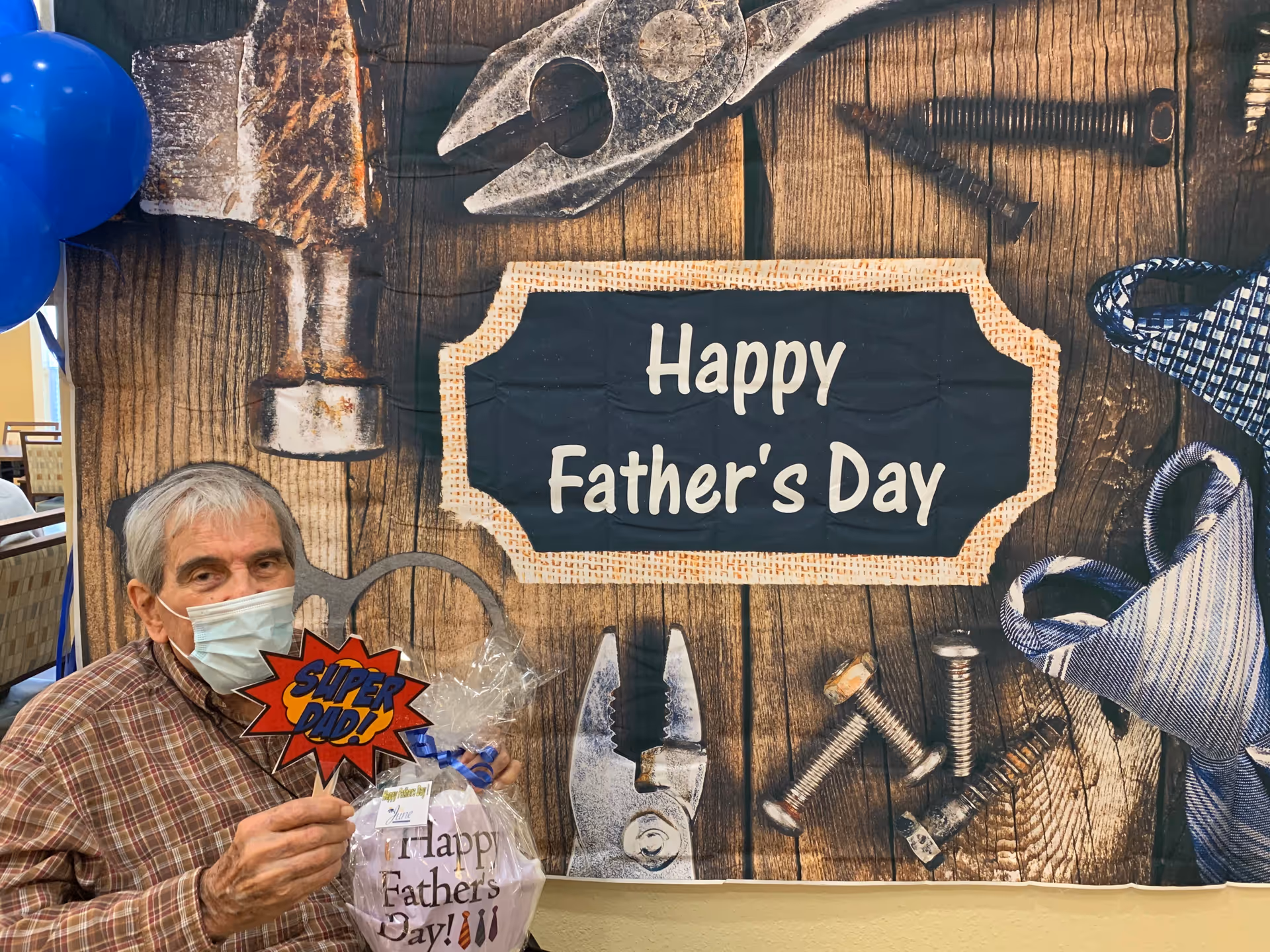 An elderly man wearing a face mask and plaid shirt holds a 'SUPER DAD!' sign and a wrapped gift in front of a wooden backdrop decorated with tools and a sign that reads 'Happy Father's Day'. Blue balloons are visible on the left side.