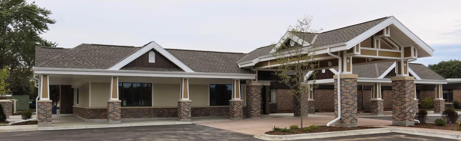 Exterior view of a single-story building with a covered entrance supported by brick columns. The building has a gabled roof with brown shingles and beige siding with brick accents. There is a small tree and some landscaping near the entrance, and a paved driveway and parking area in front.