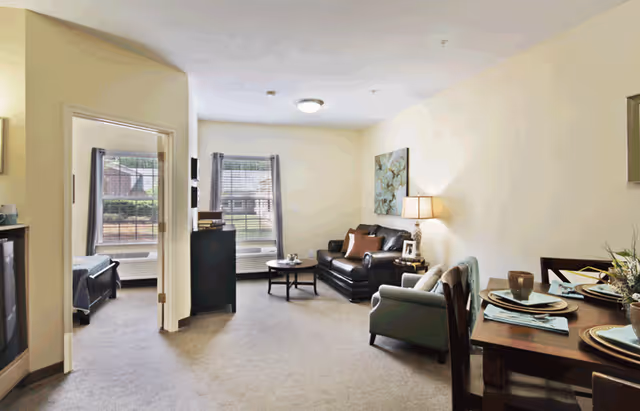 A cozy living room area with a black leather sofa, an armchair, a coffee table, and a side table with a lamp and picture frame. To the right, there is a dining table set with plates, bowls, and napkins. The room has beige walls and carpeted floors. In the background, there is a doorway leading to a bedroom with a bed and windows with blinds.