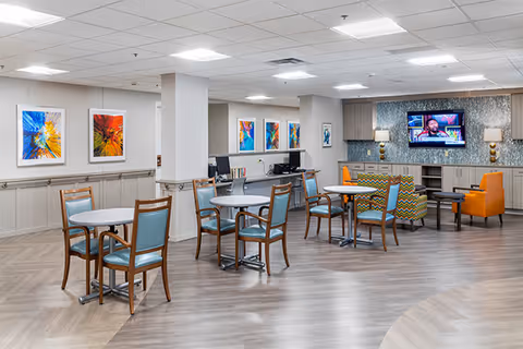 Spacious senior living common room with round dining tables and chairs, lounge seating, colorful wall art, and a wall-mounted TV.