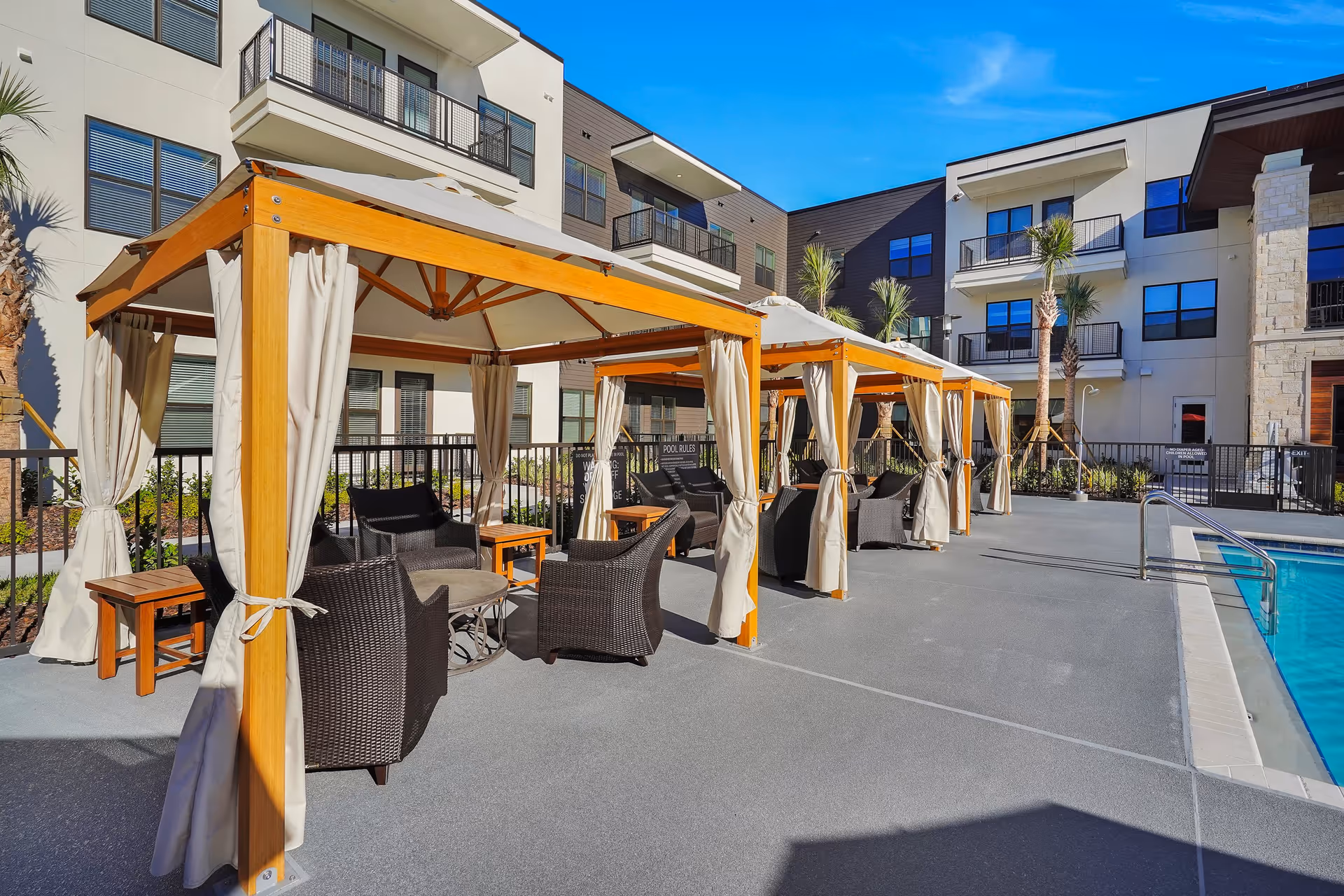 Outdoor pool area at Discovery Village Deerwood Independent Living featuring several wooden cabanas with beige curtains and wicker chairs arranged around small tables. The pool is visible on the right side, and the background shows a multi-story building with balconies and palm trees.
