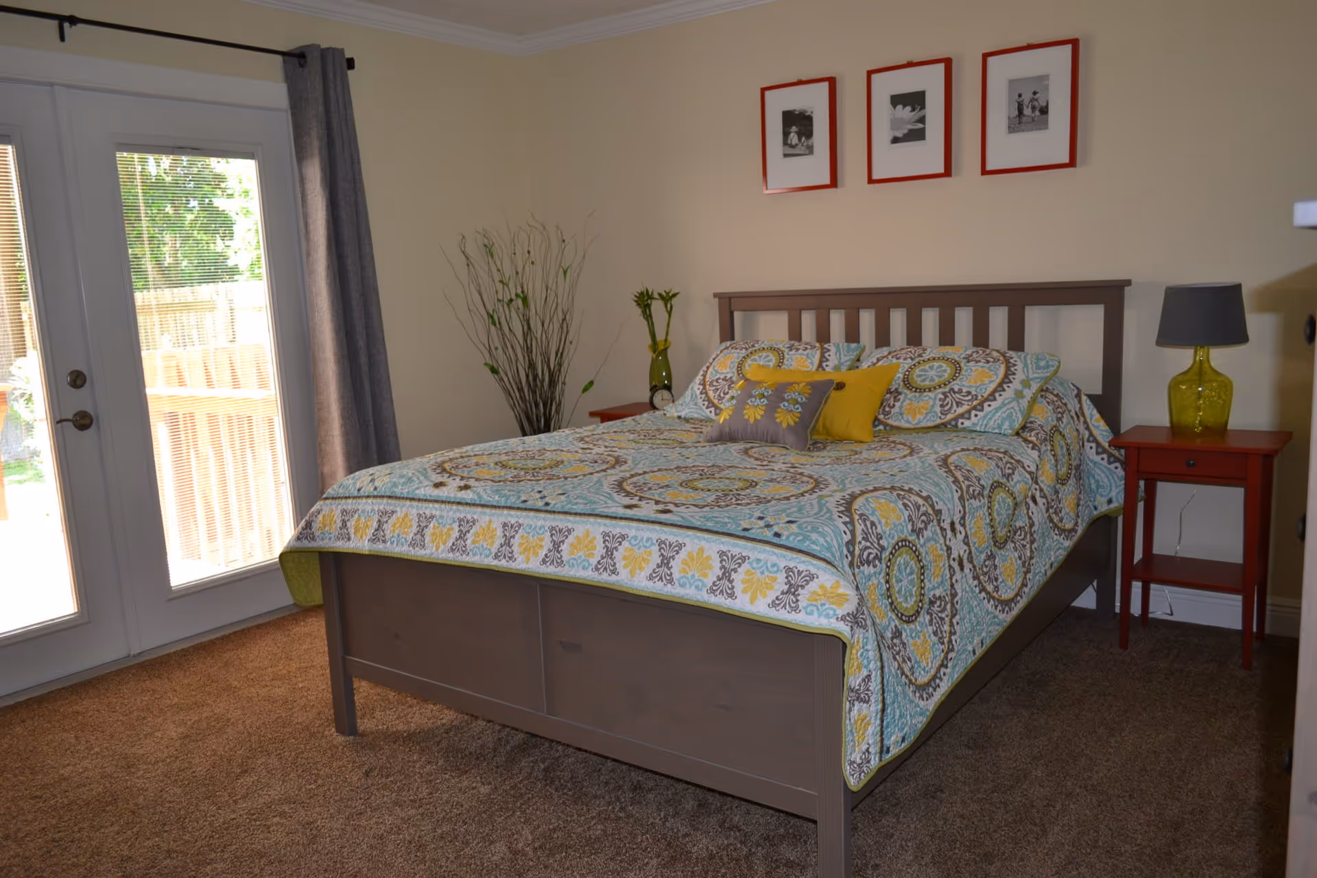 A bedroom with a wooden bed frame and a colorful patterned bedspread in shades of blue, yellow, and brown. There are three framed black and white pictures with red frames hanging on the wall above the bed. On either side of the bed are red nightstands, one with a green glass lamp and the other with a small clock and a green vase with flowers. A large window with glass doors and gray curtains is on the left side, letting in natural light. The floor is carpeted in brown.