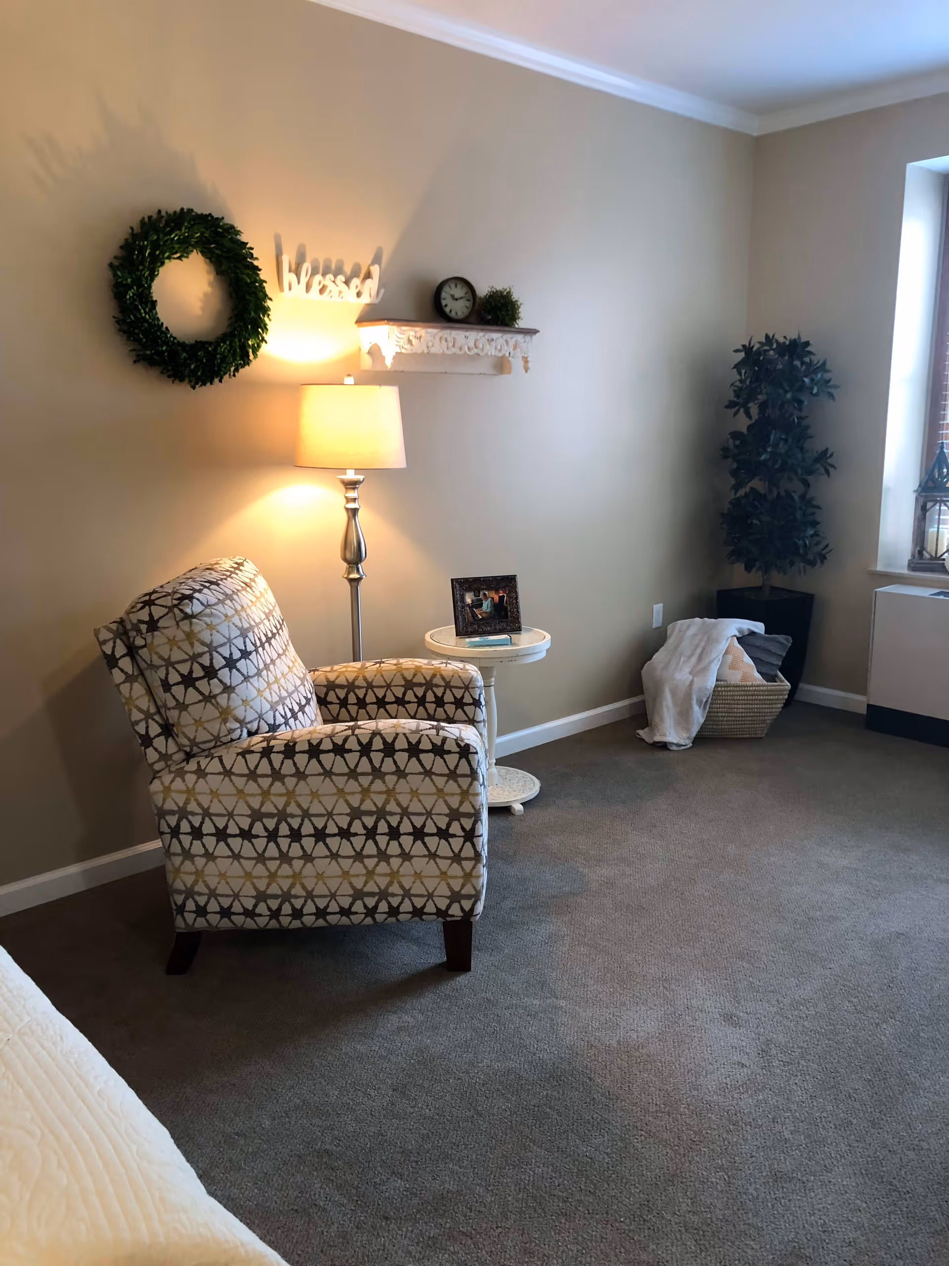 A cozy corner of a living room with a patterned armchair, a floor lamp, a small round side table with a framed photo, a decorative wreath and a shelf with a clock and small plant on the wall, a basket with blankets, and a tall potted plant near a window.