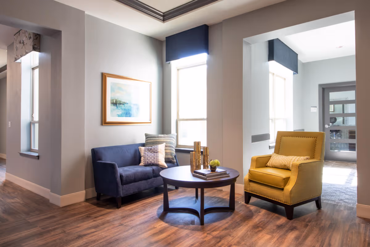 A cozy seating area in a senior living facility with a blue loveseat and a yellow armchair, both with decorative pillows. A round wooden coffee table with books and decorative items sits between the chairs. The room has light gray walls, wood flooring, and large windows with blue valances allowing natural light to fill the space. A framed abstract painting hangs on the wall above the loveseat.