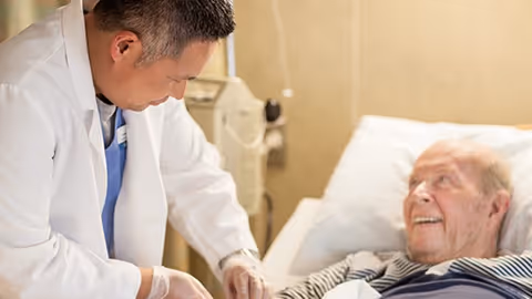 A healthcare professional in a white coat attending to an elderly male patient lying in a hospital bed, with medical equipment visible in the background.