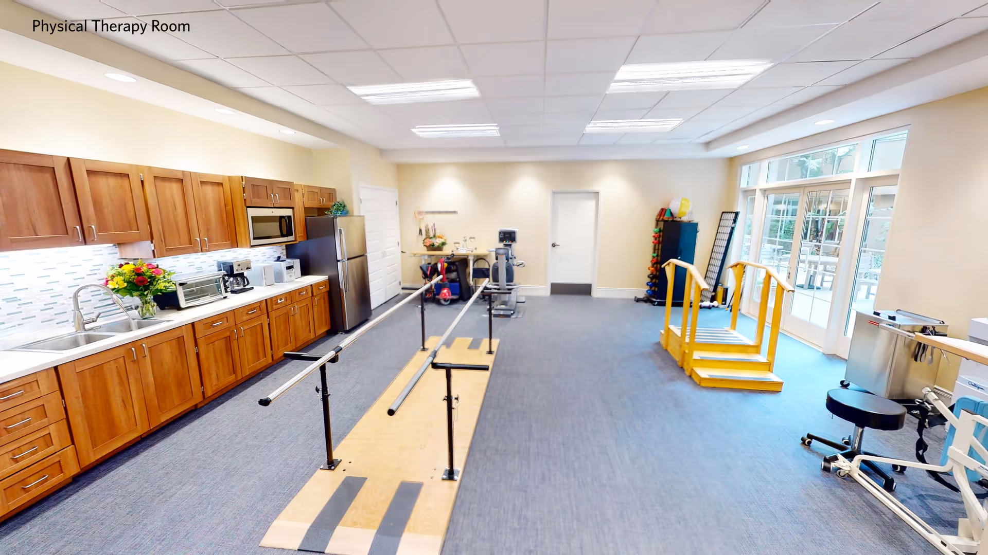 Bright physical therapy room with parallel bars, exercise equipment, a kitchenette along one wall, and large windows to the right.