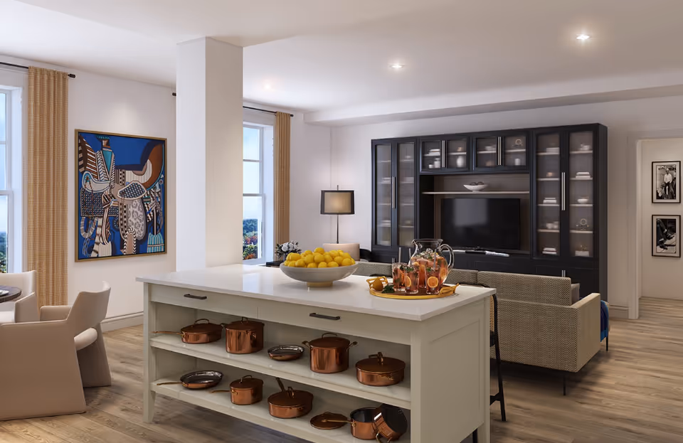 A modern open-concept living area featuring a kitchen island with copper pots and pans on shelves, a bowl of lemons, and a tray with a pitcher and glasses of iced tea. In the background, there is a beige sofa facing a black entertainment center with a TV and shelves displaying dishes. The room has large windows with beige curtains, a colorful abstract painting on the wall, and wooden flooring.