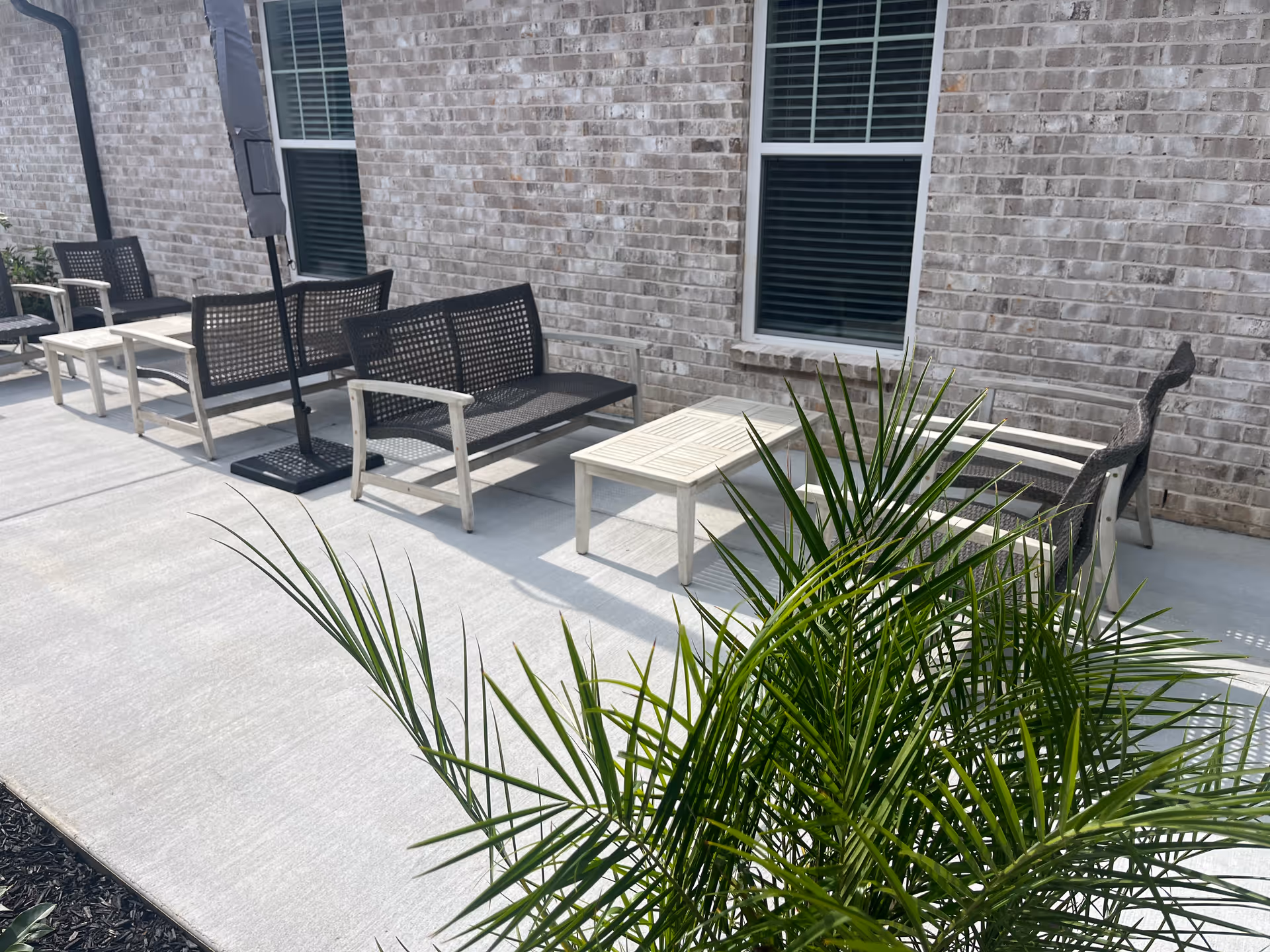 Outdoor patio area with several black and white chairs and small tables arranged along a brick wall with windows. There is a green plant in the foreground and a closed umbrella near the seating.