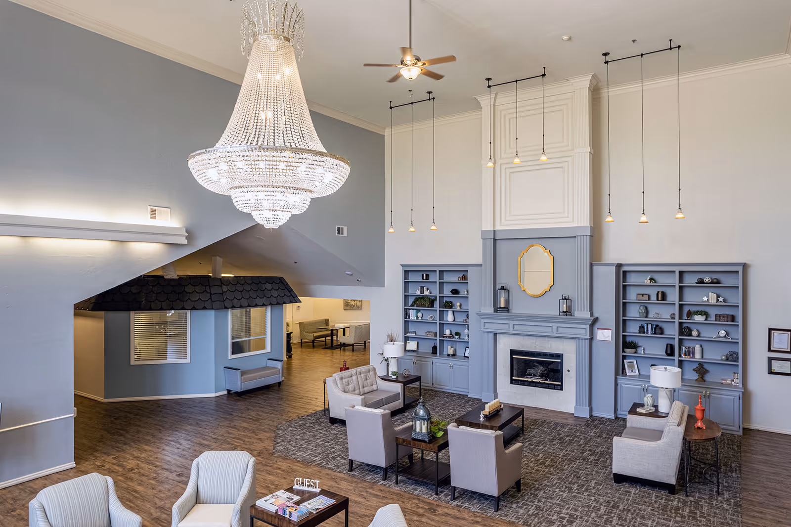Spacious senior living facility common area with high ceilings, a large crystal chandelier, ceiling fan, and multiple pendant lights. The room features a fireplace with built-in blue shelves on either side, decorated with various items. There are several comfortable armchairs and coffee tables arranged on a patterned carpet. The flooring is wood, and there is a small blue structure resembling a house with windows and a bench underneath an angled ceiling. In the background, there is a dining area with tables and chairs.