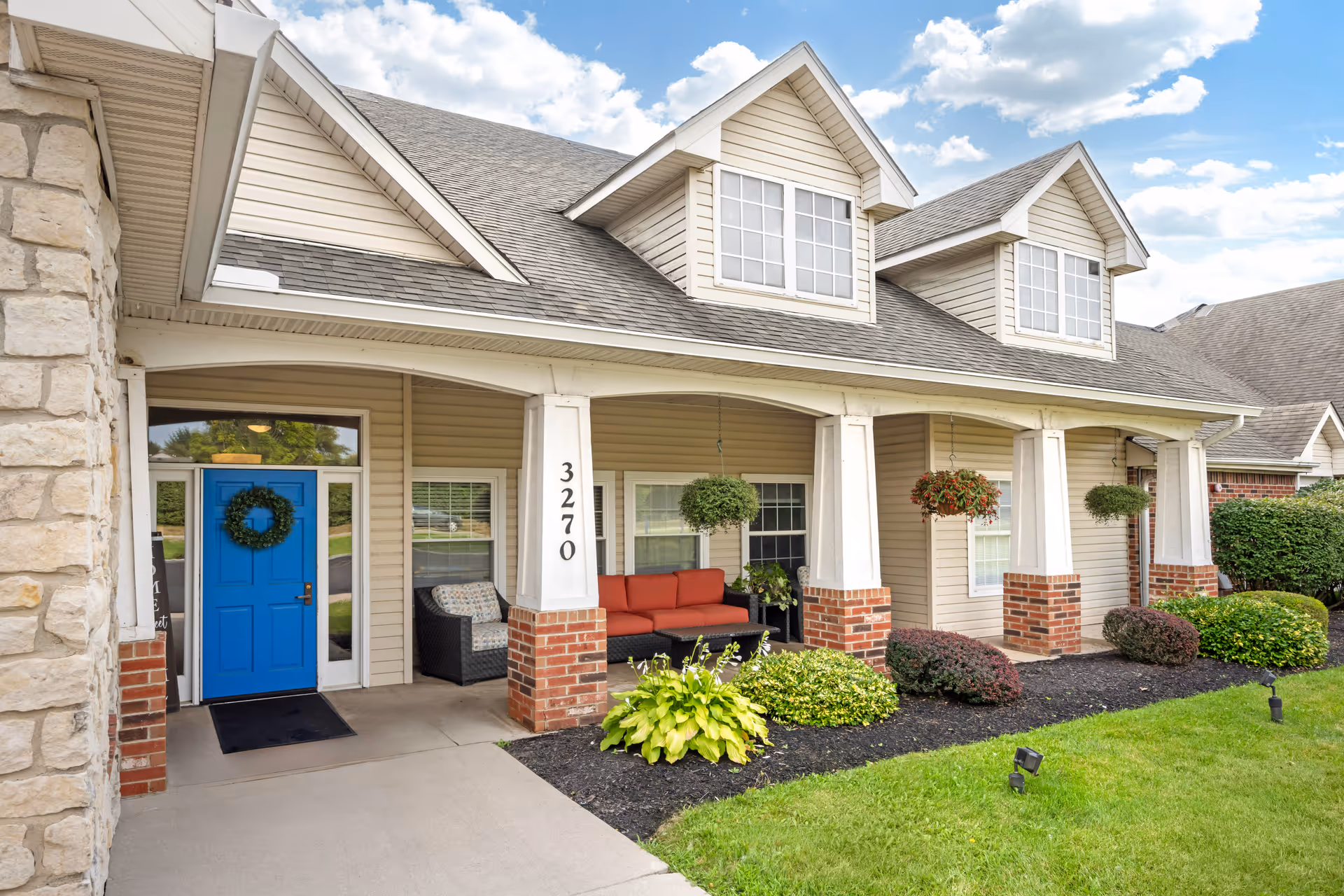 Front porch of a residential building with a bright blue door decorated with a green wreath. The porch has white columns with brick bases, hanging plants, and outdoor seating including a red cushioned sofa and a chair. The building has beige siding and dormer windows on the roof. The house number 3270 is displayed on one of the columns. There is a well-maintained garden with green shrubs and grass in front of the porch under a partly cloudy sky.