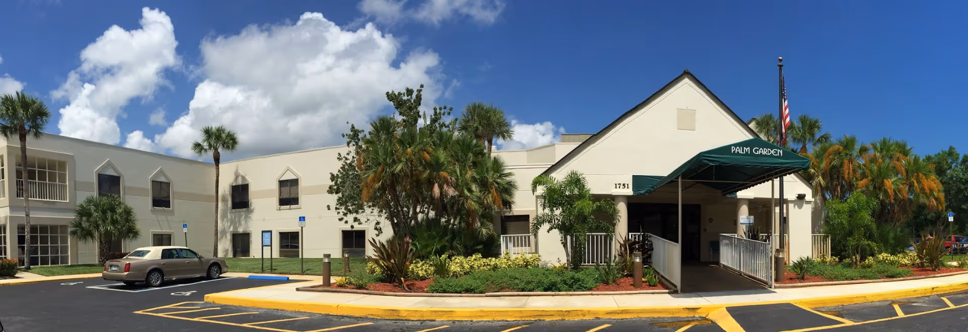 Front exterior of a senior living facility showing the entrance with a green "Palm Garden" awning, landscaped beds, palm trees, and parked cars.