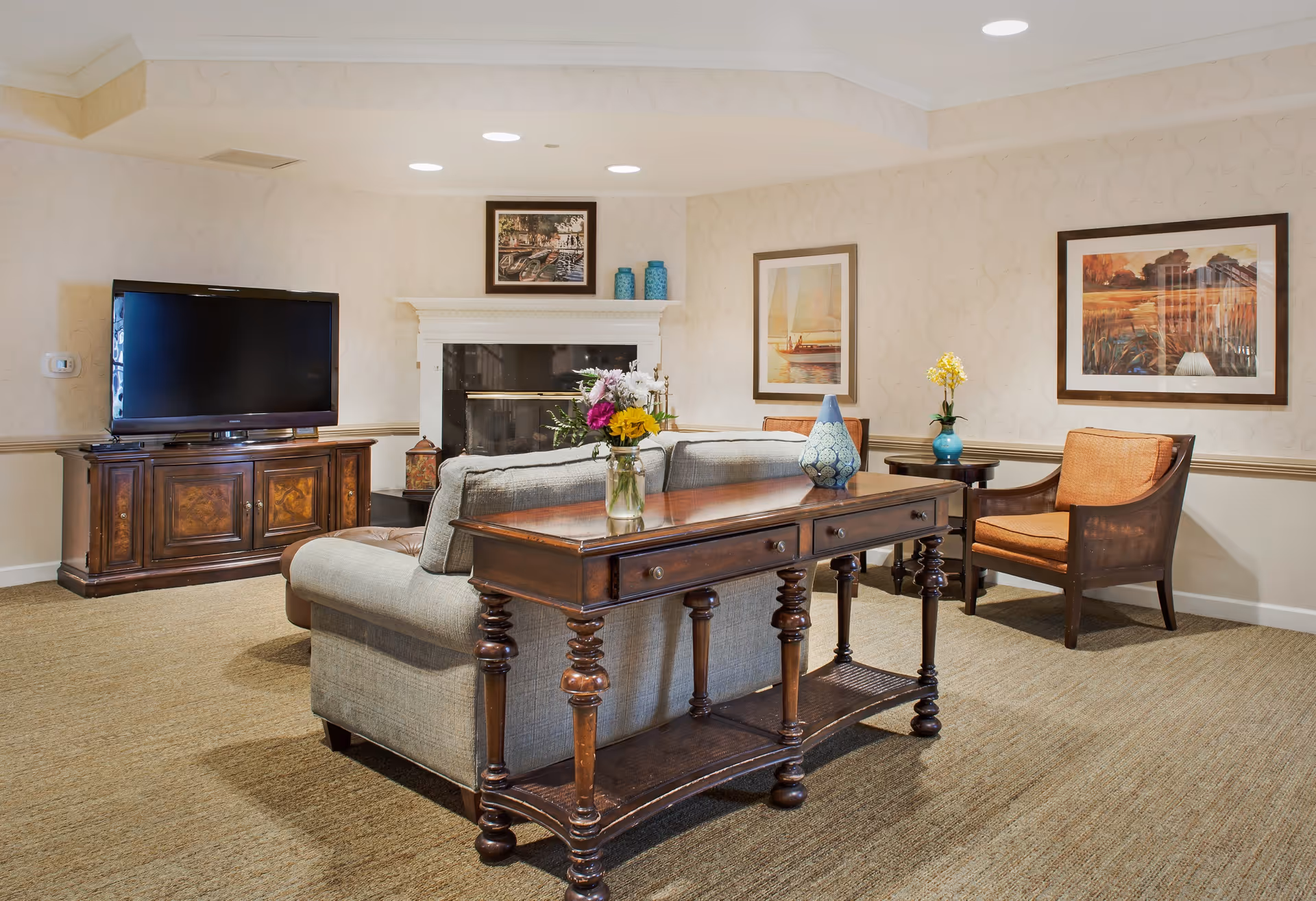 Cozy living room with a sofa and wooden console table facing a TV and fireplace, flanked by accent chairs and framed artwork.