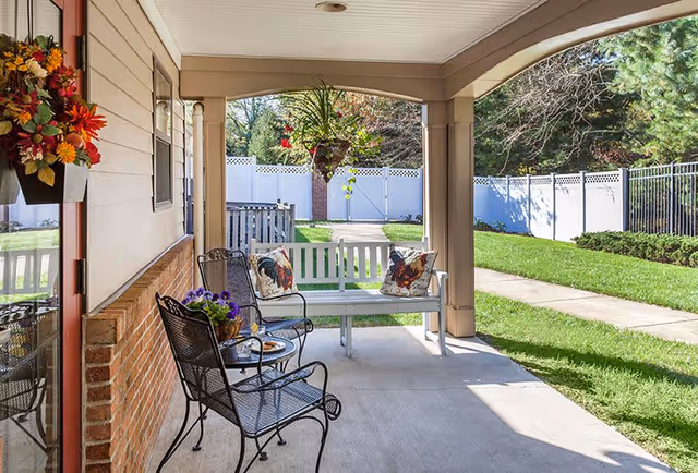 Covered outdoor patio area with a white bench adorned with two rooster-themed pillows, two black metal chairs, a small round table with a flower pot and a plate, hanging flower basket, and a view of a green lawn and white fence in the background.