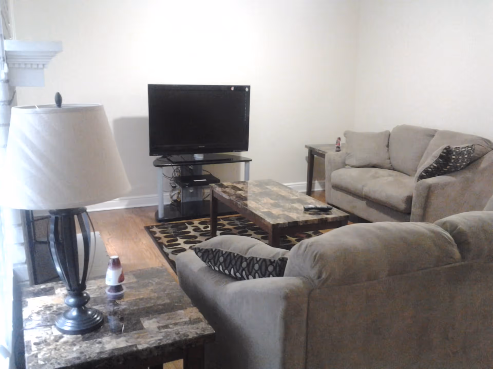 A living room with two beige sofas, a marble-patterned coffee table, a matching side table with a lamp, a flat-screen TV on a stand, and a patterned area rug on a wooden floor.
