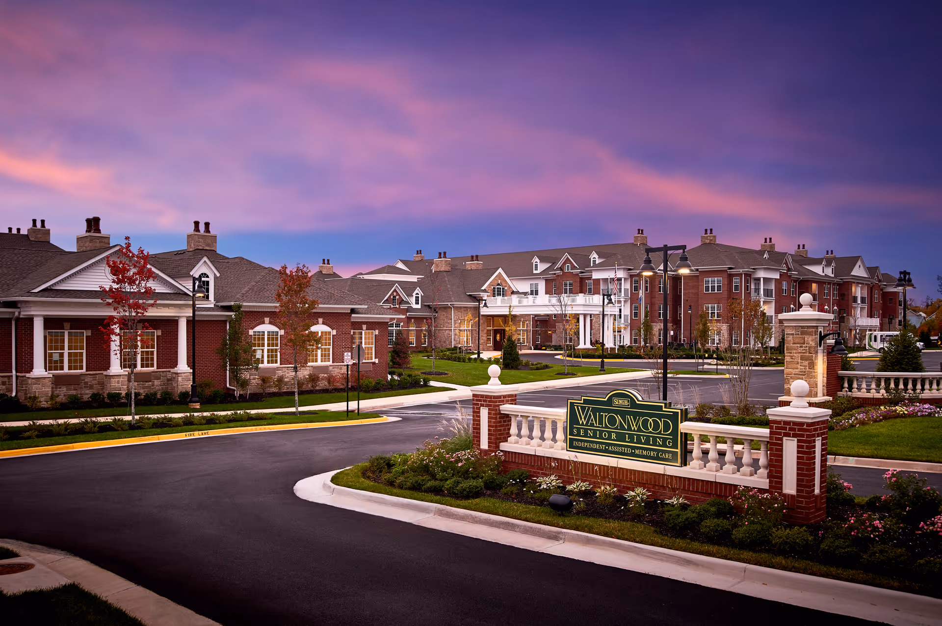Exterior view of Waltonwood Senior Living facility at dusk with a purple and pink sky. The building is large with brick and stone architecture, surrounded by landscaped greenery and a paved driveway. A sign in front reads 'Waltonwood Senior Living Independent Assisted Memory Care.'