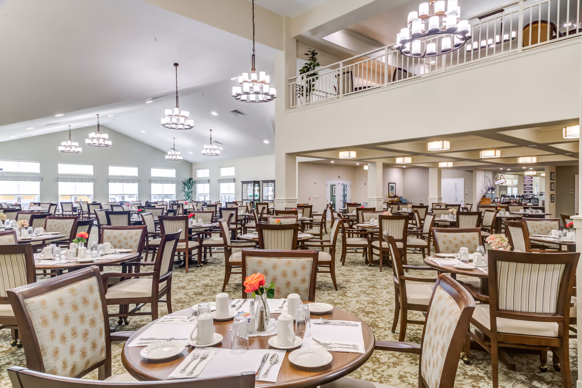 A spacious dining room with multiple round wooden tables and cushioned chairs arranged neatly. Each table is set with white placemats, plates, cups, silverware, and small flower vases with red and pink flowers. The room has high ceilings with modern chandeliers and large windows allowing natural light to fill the space. There is a second-floor balcony area visible on the right side.