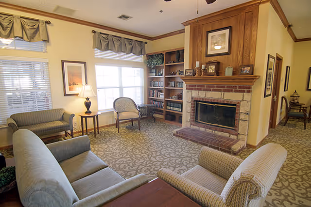 A cozy living room with patterned carpet, a brick fireplace with wooden mantel, built-in bookshelves filled with books and decorative items, several upholstered chairs and sofas arranged around a wooden coffee table, and large windows with valances letting in natural light.