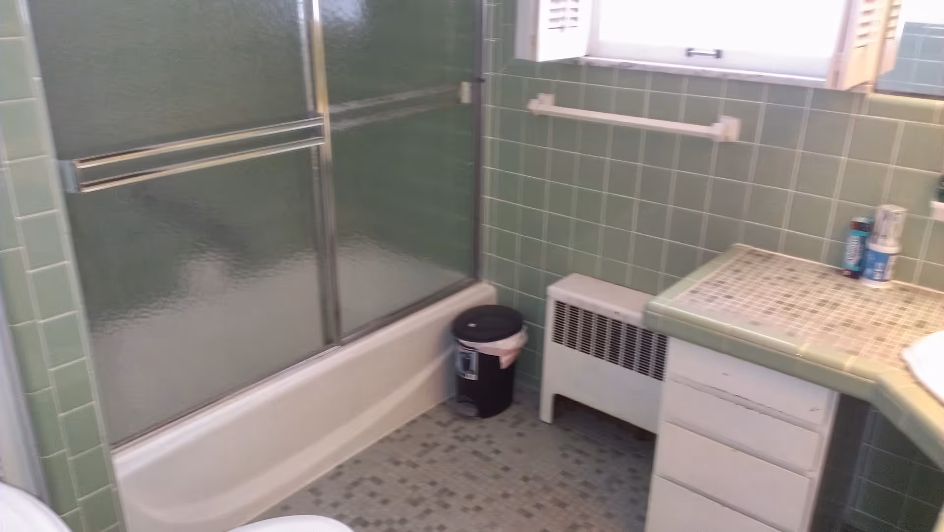 Green-tiled bathroom with a bathtub and sliding glass doors, a vanity with a mosaic countertop, heater, and small trash can.