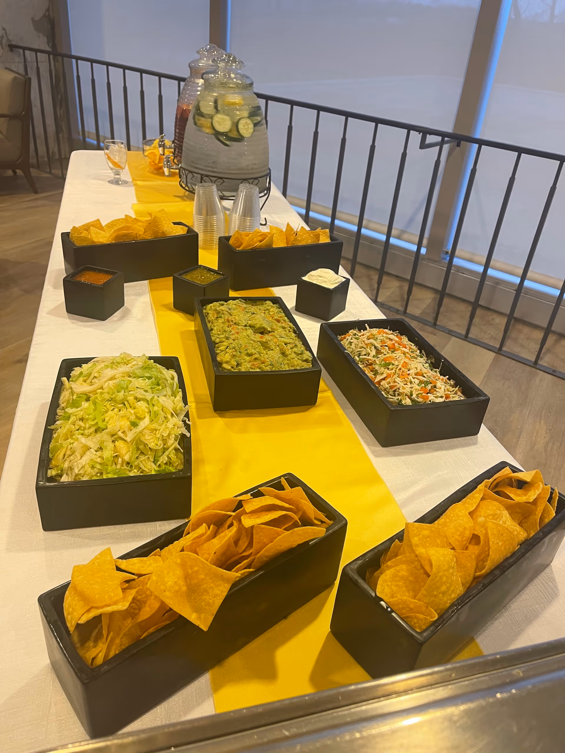A buffet table set with various foods including tortilla chips, guacamole, shredded lettuce, and a mixed salad. There are also small containers of salsa and sour cream, clear plastic cups, and a large glass beverage dispenser filled with water, lemon slices, and cucumber slices. The table is covered with a white tablecloth and a yellow runner, and is positioned near a railing with large windows in the background.