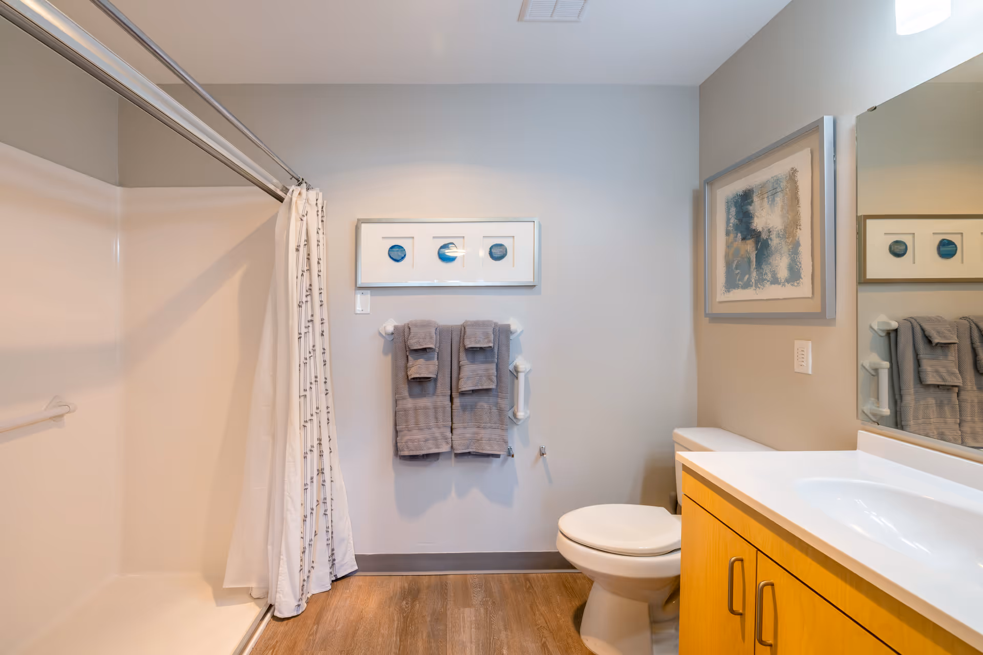 A clean bathroom with a shower area on the left featuring a white shower curtain, a toilet next to a wooden vanity with a white countertop and sink, two gray towels hanging on a towel rack on the wall, and two framed abstract artworks on the walls.