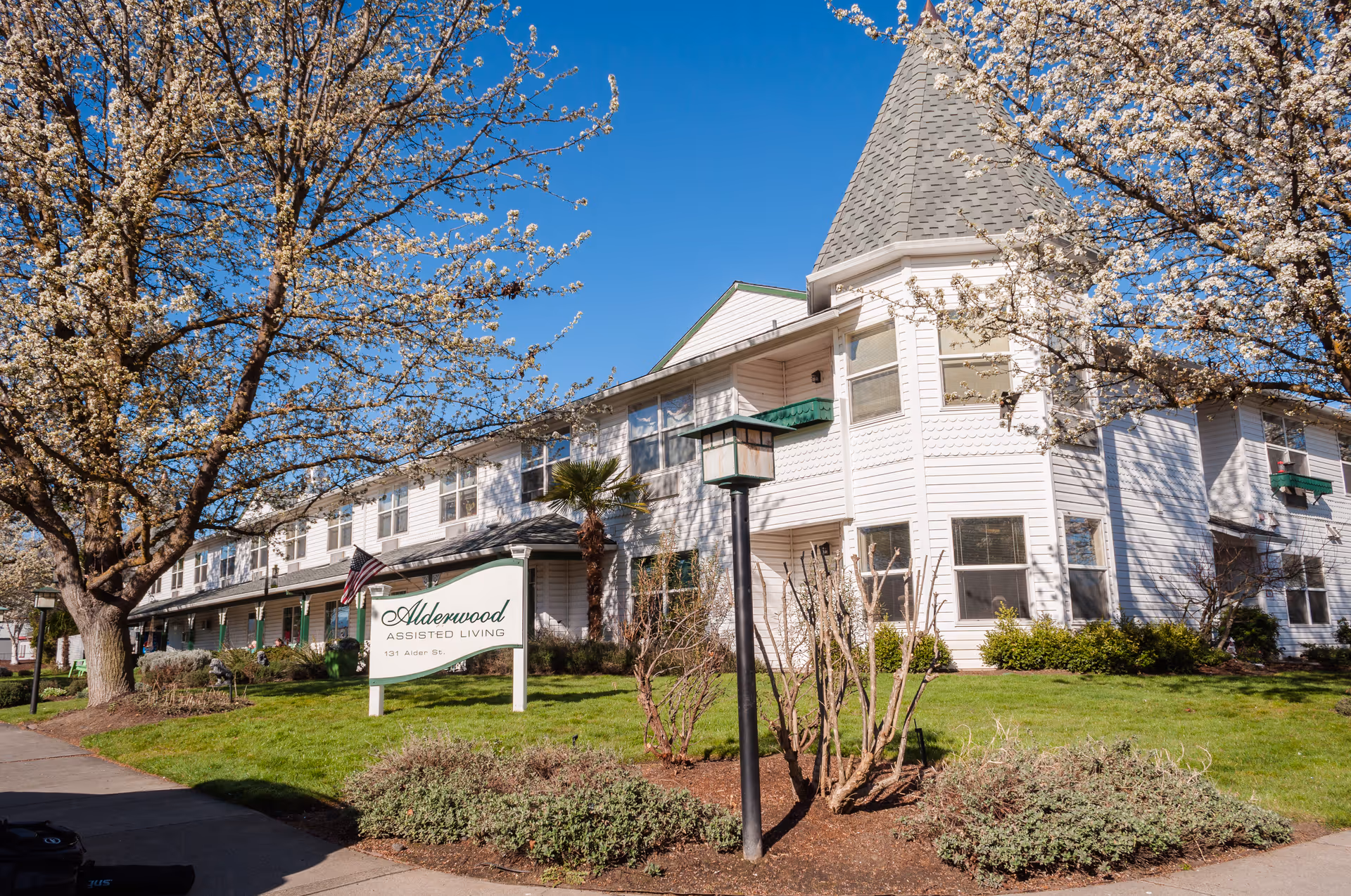 Exterior view of Alderwood Assisted Living facility with white siding, multiple windows, and a pointed roof tower. The building is surrounded by green grass, bushes, and blooming trees under a clear blue sky. A sign in front reads 'Alderwood Assisted Living, 131 Alder St.' with an American flag beside it.