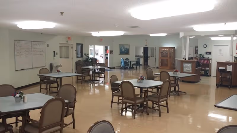 A spacious room with several square tables and chairs arranged for dining or social activities. The room has a polished floor, white walls, and fluorescent ceiling lights. There is a whiteboard on the left wall, a reception or service desk on the right, and some seating areas and doors in the background.