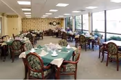 A dining room with multiple round tables covered with green tablecloths and white napkins, surrounded by floral upholstered chairs. Large windows line one side of the room, allowing natural light to fill the space. The far wall has a clock and decorative wallpaper.