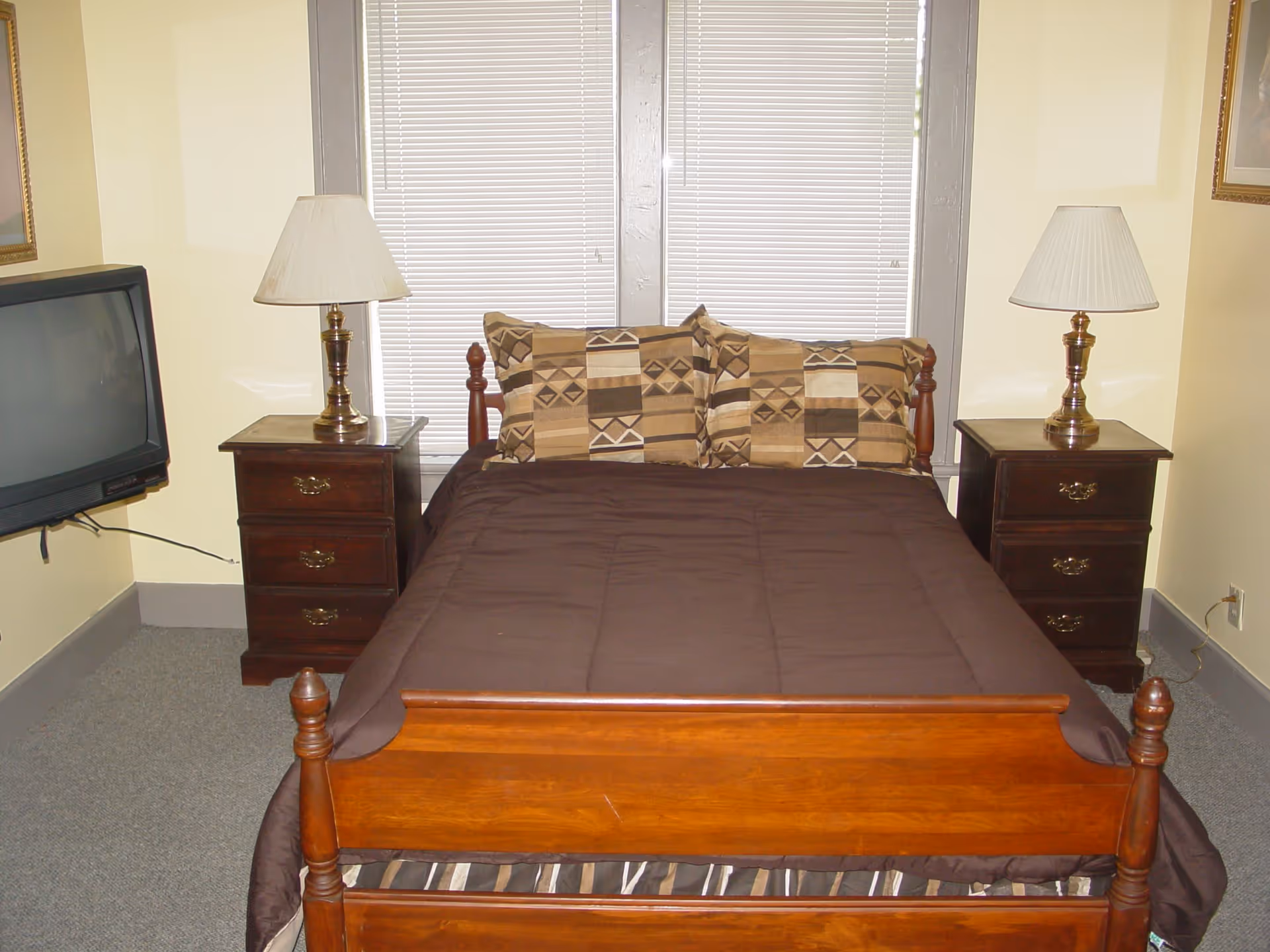 Small bedroom with a wooden bed dressed in a brown comforter, patterned pillows, two nightstands with lamps, and a wall-mounted TV.