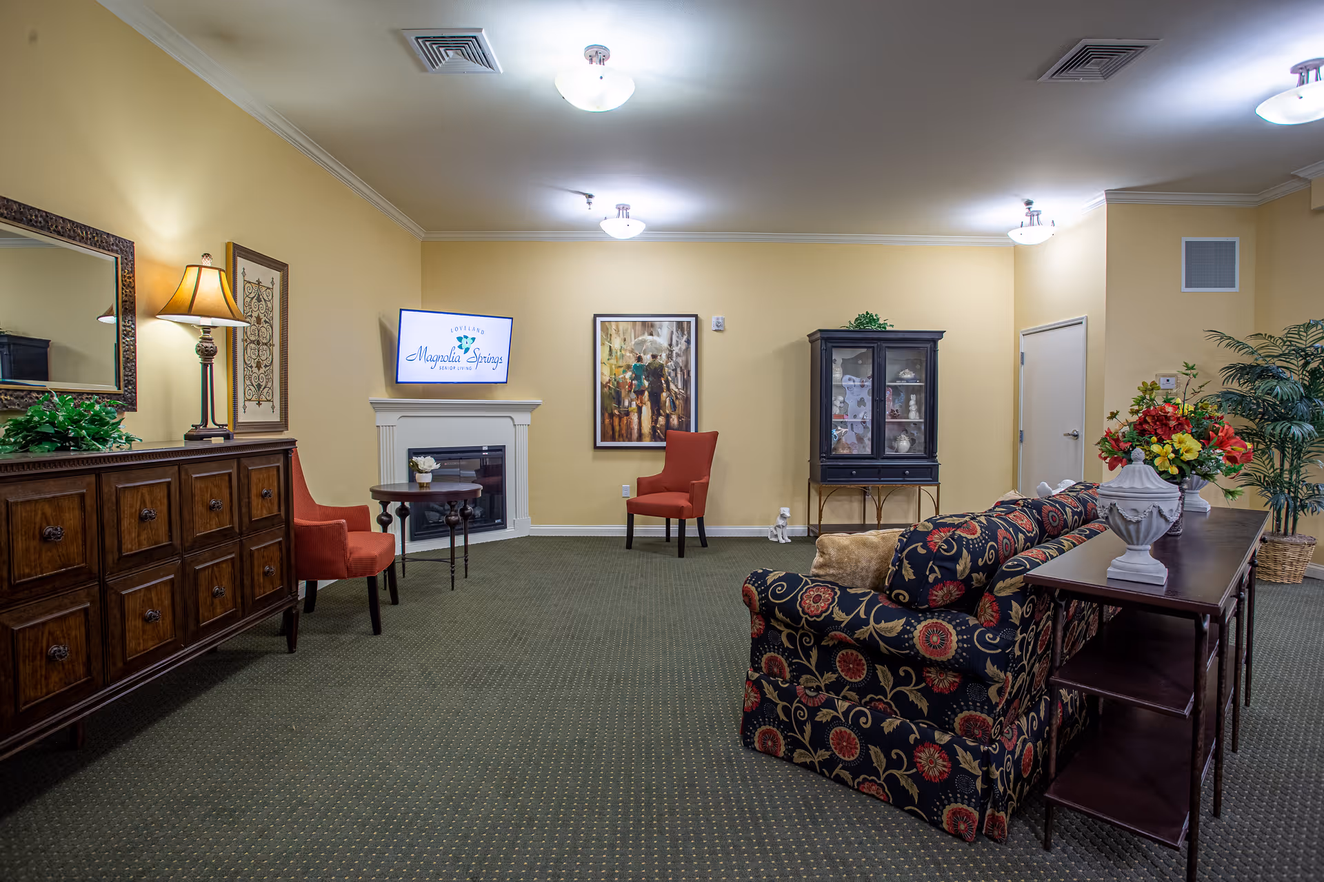 A cozy living room area with a patterned sofa, a wooden console table with a floral arrangement, two red chairs, a wooden sideboard with a lamp and a mirror above it, a small round table, a black display cabinet, and a TV mounted above a white fireplace displaying the Magnolia Springs logo. The walls are painted light yellow and the floor is carpeted in green.