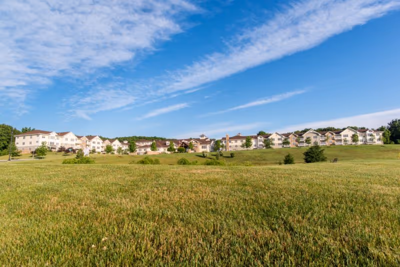 Wide view of The Heritage Of Green Hills facility buildings situated on a gentle hill with a large grassy field in the foreground under a partly cloudy blue sky.