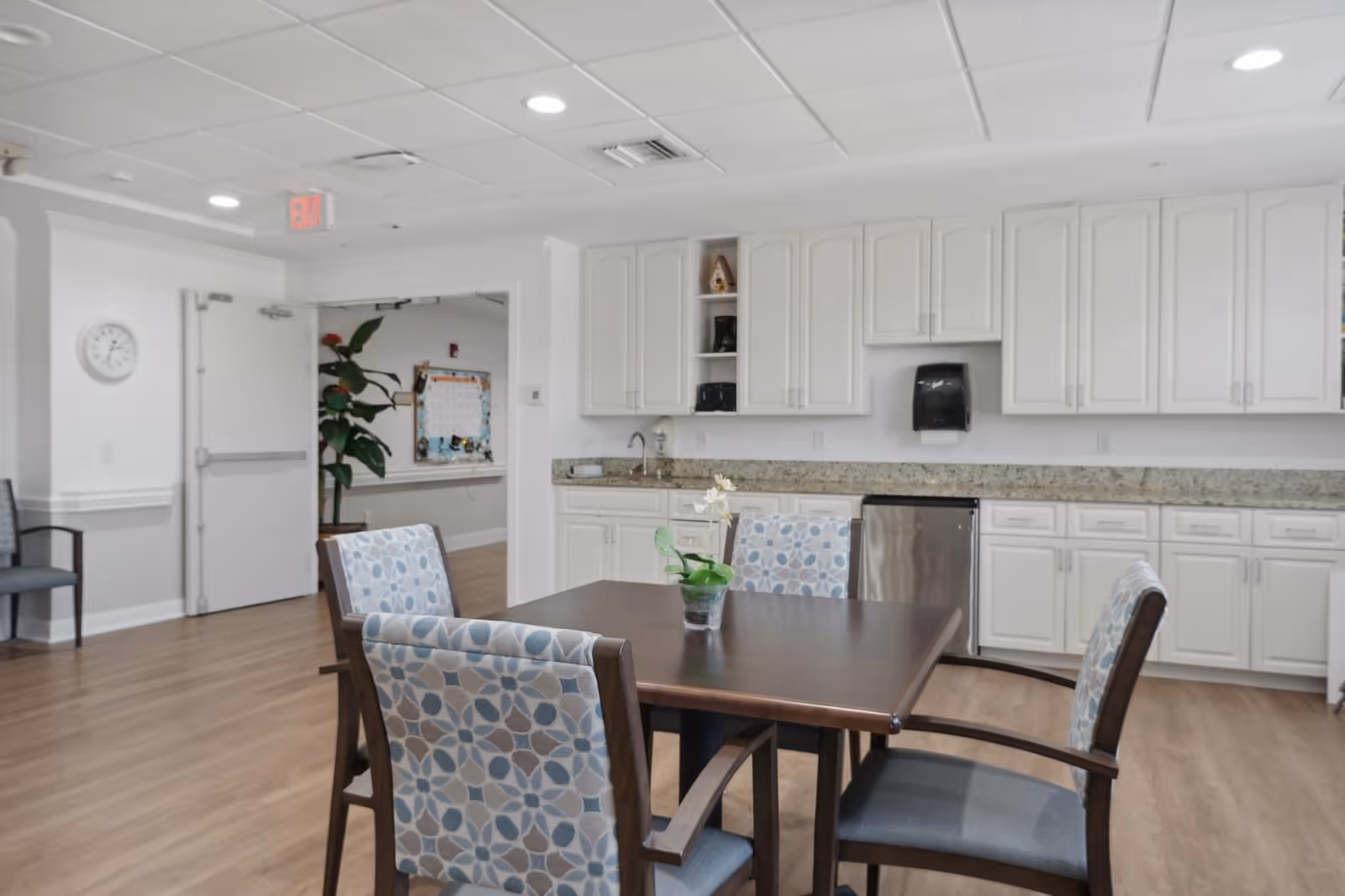 A dining room featuring a wooden table surrounded by patterned chairs, with a kitchenette in the background, including cabinets and a stainless steel refrigerator.