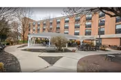 Outdoor patio area with tables and chairs under a large white canopy, situated in front of a multi-story brick building with many windows. The area is surrounded by paved walkways and some trees with bare branches.