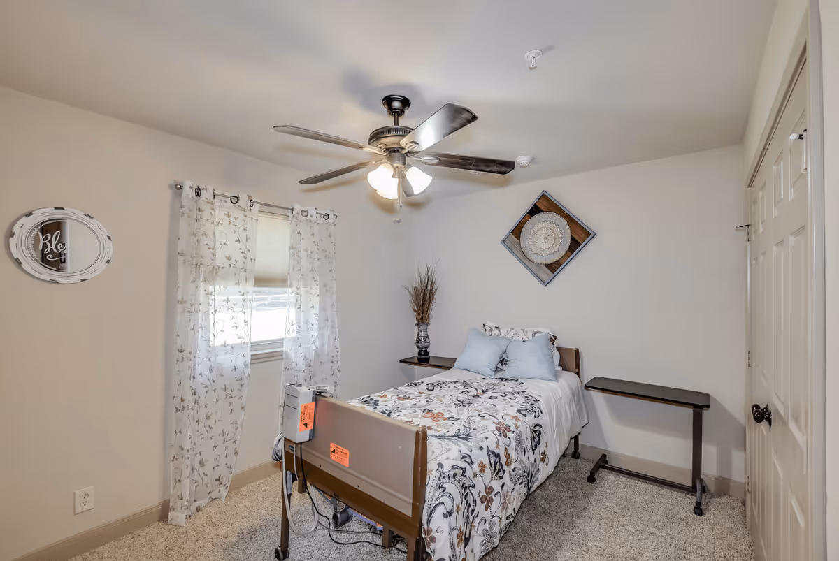 A small bedroom with a single bed featuring floral bedding and two light blue pillows. The bed has medical equipment attached to its footboard. The room has beige walls, a ceiling fan with lights, a window with sheer floral curtains, a decorative wall piece above the bed, a vase with dried plants on a small shelf, and a small overbed table next to the bed. There is a closed door on the right side of the room.