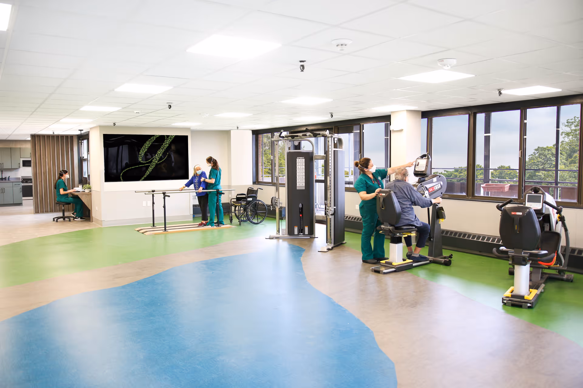 A spacious rehabilitation and exercise room with large windows letting in natural light. Two healthcare workers in green scrubs assist elderly patients using exercise equipment and parallel bars. One patient is seated on a stationary exercise bike, while another is walking with support. A wheelchair is visible near the parallel bars. The floor has a colorful design with green, blue, and beige sections. In the background, a staff member is seated at a desk working on a computer.