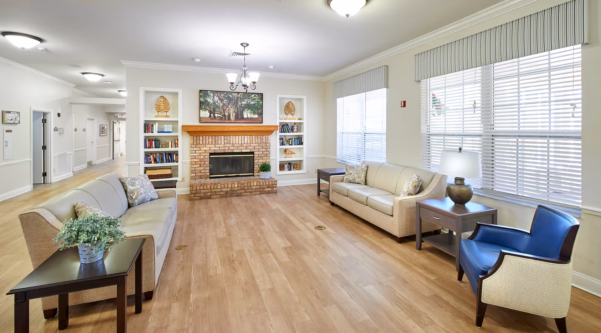 A bright and spacious living room in a senior living facility with two beige sofas, a blue armchair, wooden side tables with lamps and plants, a brick fireplace with a wooden mantle, built-in shelves with books and decorative items, large windows with blinds, and light wood flooring.