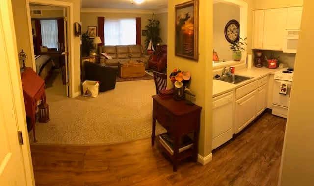 View of a cozy living area and kitchen in a senior living facility. The living room features a beige sofa, a black armchair, a wooden coffee table, and a large window with curtains. The kitchen has white cabinets, a sink, a stove, and a dishwasher. A small wooden table with flowers and a painting on the wall separates the kitchen from the living area. A bedroom is visible through an open door on the left side.