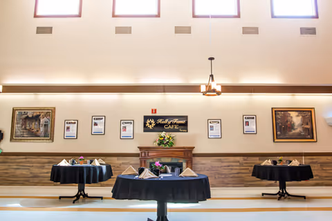 Interior view of a dining area with three round tables covered with black tablecloths, each set with napkins and small flower arrangements. The room has high windows near the ceiling letting in natural light, a chandelier hanging from the ceiling, framed pictures and a sign reading 'Hall of Fame Cafe' on the wall above a wooden mantel.