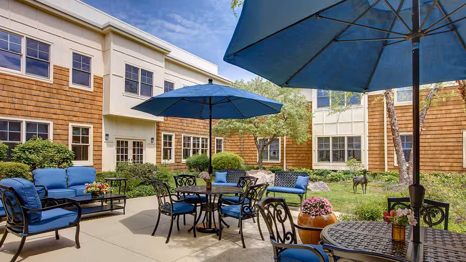 Outdoor patio area at a senior living facility with blue cushioned chairs and sofas, round tables with blue umbrellas, potted flowers, and greenery surrounding the space. The building exterior features brown shingles and white trim with multiple windows.