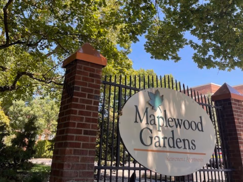Entrance gate of Maplewood Gardens retirement apartments with a large oval sign mounted on black iron fencing, flanked by brick pillars topped with copper caps, surrounded by green trees under a clear blue sky.