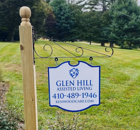 A white and blue sign hanging from a wooden post on a grassy lawn. The sign reads 'Glen Hill Assisted Living 410-489-1946 kenwoodcare.com' with a logo above the text. Trees and a green landscape are visible in the background.
