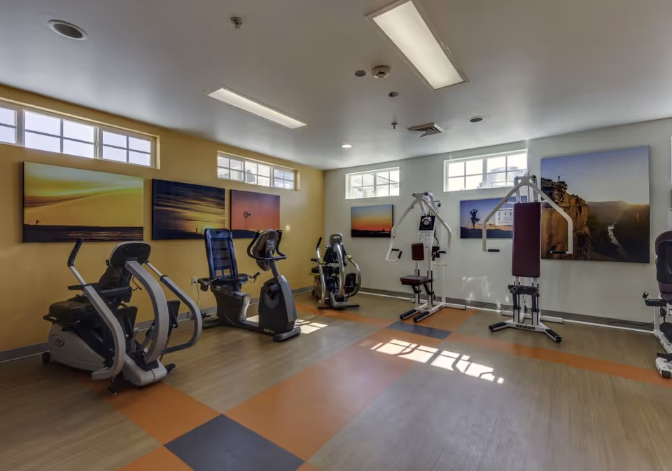 A well-lit exercise room with various fitness equipment including stationary bikes and weight machines. The room has large windows near the ceiling allowing natural light to enter. The walls are decorated with large landscape photographs featuring sunsets and nature scenes. The floor has a patterned design with orange, gray, and beige tiles.