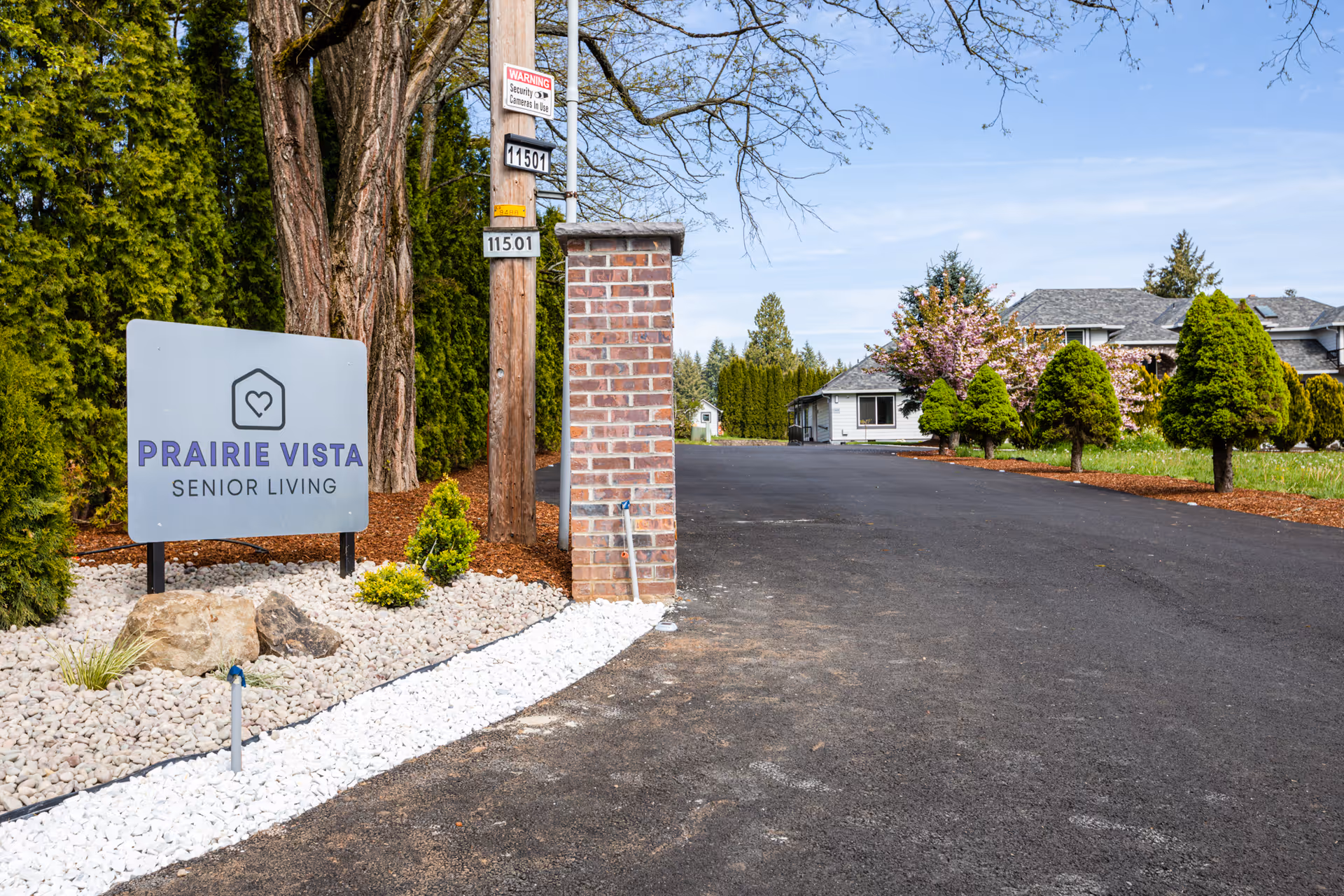 Entrance to Prairie Vista Senior Living facility with a sign displaying the facility name, a paved driveway, landscaped area with rocks and small shrubs, tall trees, and a building in the background under a clear sky.