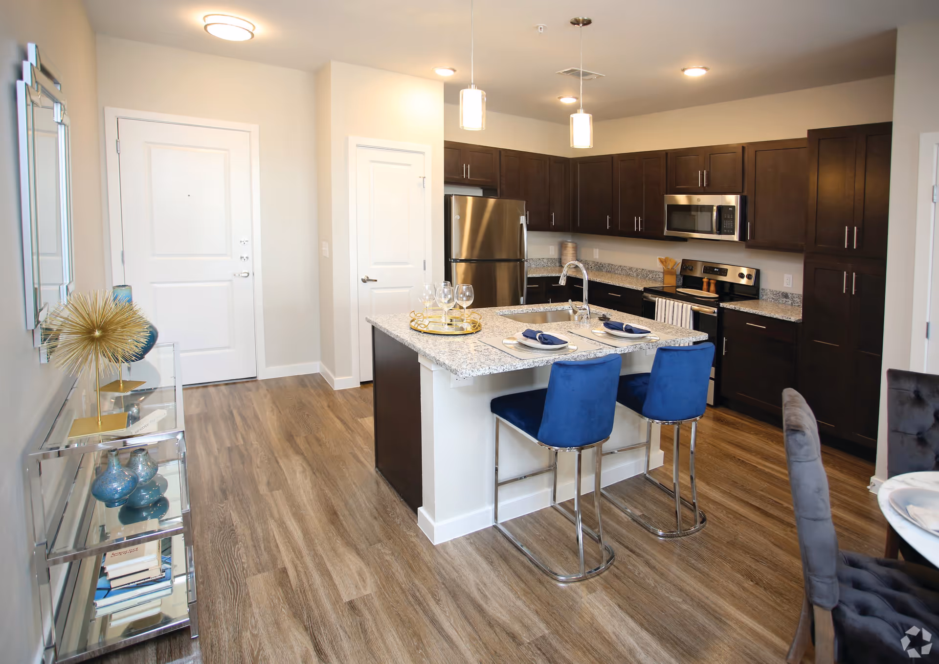 Modern kitchen with dark wood cabinets, stainless steel refrigerator, microwave, and stove. A kitchen island with a granite countertop has a sink and two blue bar stools. The floor is wood, and there is a glass shelving unit with decorative items on the left side near the entrance door.