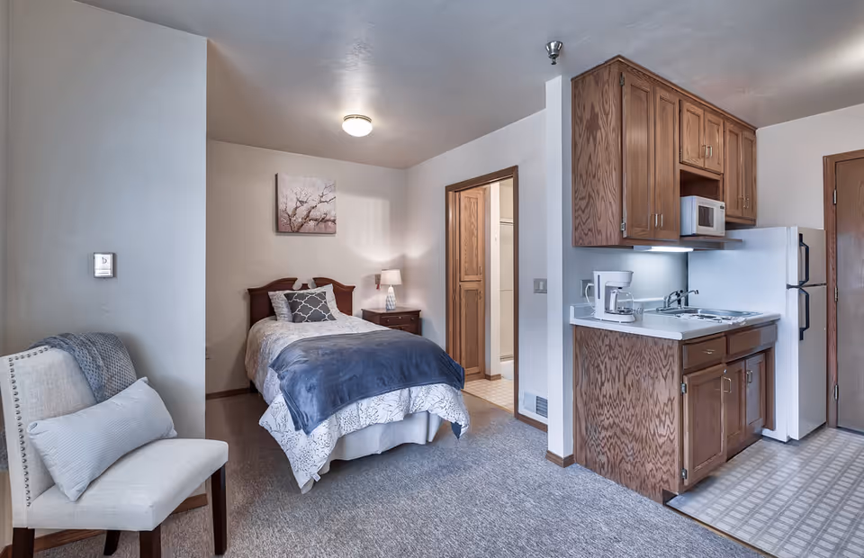 A cozy studio apartment featuring a bed with a blue blanket and decorative pillows, a wooden nightstand with a lamp, a white cushioned chair with a pillow and throw blanket, and a compact kitchen area with wooden cabinets, a microwave, a coffee maker, a sink, and a refrigerator. The room has carpeted flooring and a door leading to another room or bathroom.