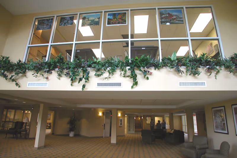 Interior view of a senior living facility lobby area with seating arrangements, carpeted floor, and large windows on the upper level decorated with framed pictures and green plants along the ledge.