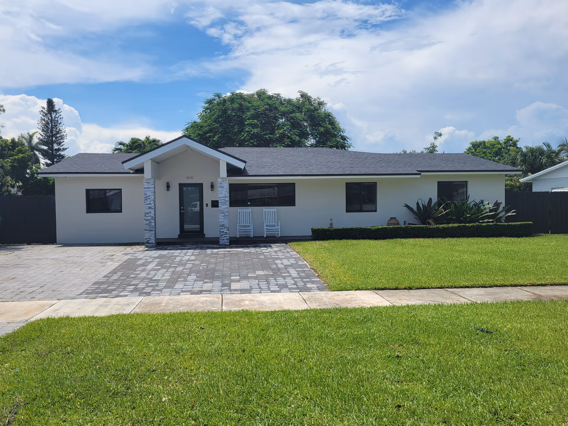 Front exterior of a single-story white house with a paved driveway, green lawn, and two white rocking chairs on the porch.