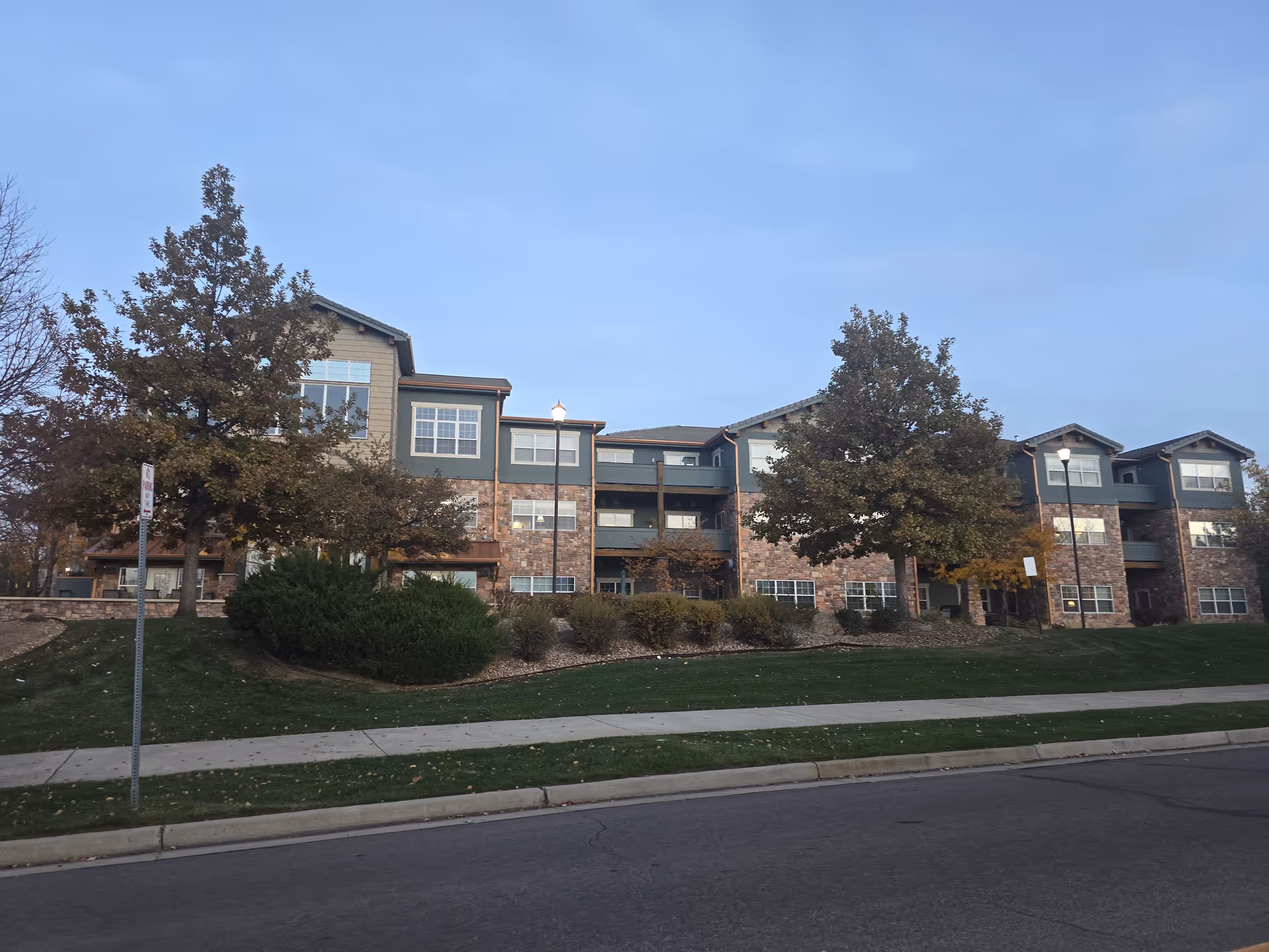 Front exterior of a three-story assisted living building with brick and siding, trees and a sidewalk in the foreground under a clear sky.